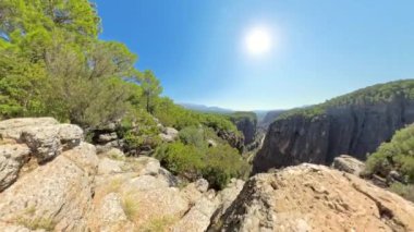 Bir kadın derin bir kanyona bakan ve yemyeşil ormanlarla çevrili kayalık bir uçurumda duruyor. Geniş açı, dik düşüşü ve geniş manzarayı vurguluyor..