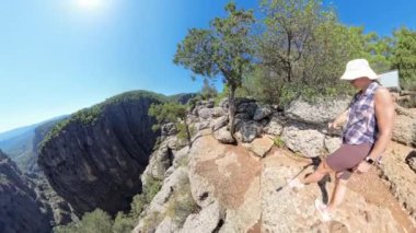 Bir kadın, arka planda Kanyon ve Dağlar 'ı dramatik bir şekilde gören bir uçurumun kenarında yatıyor. Geniş açılı çekim, parlak güneşin altında engebeli kayaları ve ağaçlarla kaplı yolları yakalar..