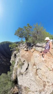 Dikey video. Bir kadın, arka planda Kanyon ve Dağlar 'ı dramatik bir şekilde gören bir uçurumun kenarında yatıyor. Geniş açılı çekim, parlak güneşin altında engebeli kayaları ve ağaçlarla kaplı yolları yakalar..