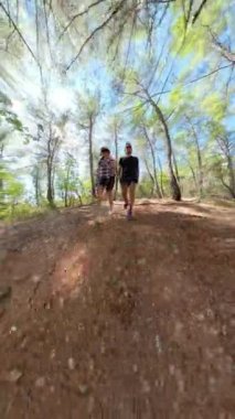 Dikey video. Küçük gezegen. Bir adam ve bir kadın, alçak bir perspektiften yakalanarak orman yolunda yürür. Geniş açı, engebeli araziyi ve parlak güneş ışığı altında yükselen ağaçları vurguluyor..