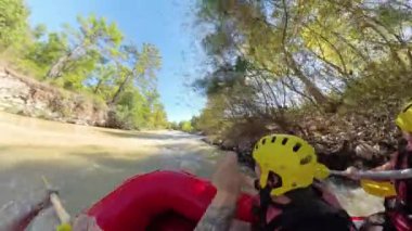 Bir rafting teknesinin içinde, her iki taraftaki ağaçlarla çevrili ve güvenlik için sarı miğferler takan ekip nehrin sakin bir kısmında kürek çekerken yan açılı bir görüntü..