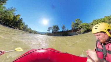 Bir rafting teknesinin ön tarafından çekilen alçak açılı bir çekim. Sarı başlıklı iki kirişin parlak bir güneşin altında kayalıklarla ve yeşilliklerle çevrili bir nehirde kürek çekmesi..