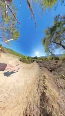 Dikey video. Ormanda kayarak ilerleyen bir kadının dinamik bir görüntüsü. Arka planda açık gökyüzü ve dağ manzarası var. Güvenlik için kask ve emniyet kemeri takıyor..