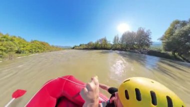 Geniş bir nehirde kürek çeken bir grup kirişin yan açılı görüntüsü, sarı kasklar ve can yelekleriyle nehir kıyısı boyunca ağaçlar ve parlak bir güneş tepemizde parlıyor..