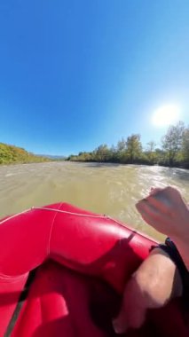 Dikey video. Geniş bir nehirde kürek çeken bir grup kirişin yan açılı görüntüsü, sarı kasklar ve can yelekleriyle nehir kıyısı boyunca ağaçlar ve parlak bir güneş tepemizde parlıyor..