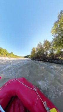 Dikey video. Ağaçlarla çevrili hızlı hareket eden nehir bölümüne yaklaşan bir rafting teknesinin ön tarafındaki görüntüde güneş ışığı yaprakların arasından süzülüyor ve sarı kürekler görülebilir..