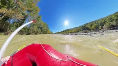 Akıntıya karşı giden bir salın alçak açılı görüntüsü, hareket halindeki küreklerle ve nehir kıyısında uzanan ağaçlarla, parlak güneşin altında macera ve dinamik bir hareket hissi uyandırıyor..