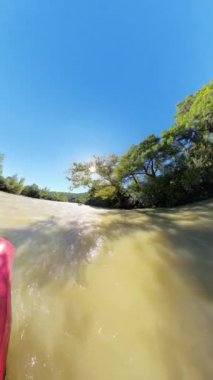 Dikey video. Dikey video. Çamurlu bir nehirde, parlak bir güneşin altında, sudan yansıyan ağaçlık kıyılar ve güneş ışığı ile birlikte, hareketli bir hareket ve açık hava macerası hissi yaratan, kırmızı bir salın alçak açılı çekimi..