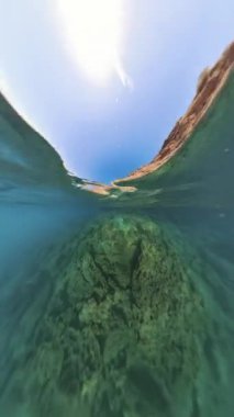 Dikey video. Suyun yüzeyindeki kayalık kayalıkların yansımasını yansıtan barışçıl bir sualtı görüntüsü, sualtı manzarasını gösteren turkuaz bir su..