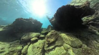 Güneşli kayalar ve berrak mavi suyla çevrili bir insan su altında yüzüyor. Güneş ışığı aşağı akıyor, sakin ve huzurlu bir manzara yaratıyor. Çekim, yüzücüyü 360 derecelik bir açıyla yakalıyor..