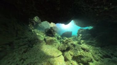 Suyun içinden geçen ve kayalar ve mercanlarla çevrili bir sualtı mağara girişini aydınlatan çarpıcı bir su altı görüntüsü. Manzara mağaranın ötesindeki derin mavi denize bakıyor..