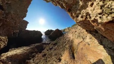 360 derece dönüş. Güneş ışığı, pürüzlü kayaların sakin denizle buluştuğu kayalık bir kıyı manzarasının üzerinde parlar. Engebeli kıyı şeridi ve parlak gökyüzü huzurlu, el değmemiş bir deniz manzarası oluşturuyor..