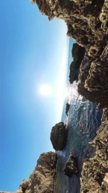 Dikey video. 360 dönüş, yakınlaştır. Kayalık bir yarıktan bakarken, parlak güneş uzaktan denizden yansıyor. Sivri kayalar ve berrak mavi gökyüzü dramatik ve çarpıcı bir kıyı manzarası yaratır..
