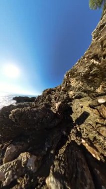 Dikey video. 360 derece dönüş. Kayalık bir geçit, güneşin denizin üzerinde parladığı parlak bir kıyı manzarasına açılıyor. Engebeli kaya oluşumları ve sakin su manzaralı bir deniz kenarı oluşturur..