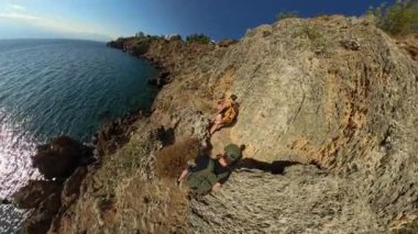 İki yürüyüşçü engebeli bir sahil yolunda yürürken güneş denizden yansıyor. Kayalık uçurumlar, berrak gökyüzü ve okyanus manzarası açık hava keşiflerinin güzelliğini vurgular..