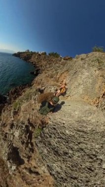 Dikey video. İki yürüyüşçü engebeli bir sahil yolunda yürürken güneş denizden yansıyor. Kayalık uçurumlar, berrak gökyüzü ve okyanus manzarası açık hava keşiflerinin güzelliğini vurgular..