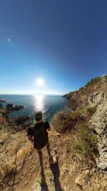 Dikey video. İki yürüyüşçü, okyanusa bakan dar ve dik bir patika boyunca ilerler. Güneş ışığı suda parlarken, kayalık uçurumlar kıyı şeridinin engebeli güzelliğini artırıyor..