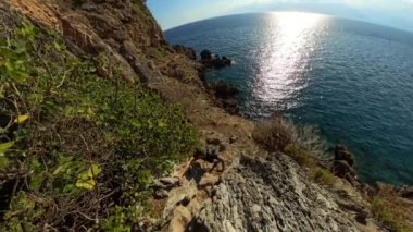 Okyanus yakınlarındaki bir uçuruma tırmanan bir dağcıyla dik bir sahil tırmanışı. Güneş ışığı aşağıdaki denizden yansıyarak engebeli araziyi ve dramatik manzarayı vurgular..