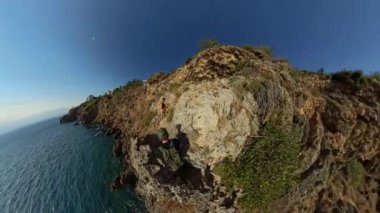 Dağcıların kayalık bir sahil uçurumuna tırmanırken geniş açılı bir manzarası var ve okyanus aşağıya doğru uzanıyor. Dramatik yükseklik ve açık gökyüzü yolculuğun maceralı doğasını vurgular..