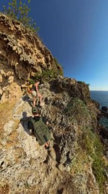 Dikey video. Dik kayalık bir çıkıntıya tırmanan dağcılarla dolu bir sahil macerası. Aşağıdaki parlak güneş ve parıldayan okyanus yükseklik ve engebeli bir keşif hissi yaratıyor..