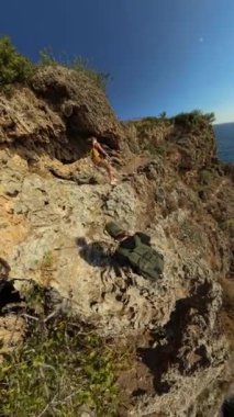 Dikey video. Yürüyüşçüler, aşağıdaki okyanusa yansıyan güneşle dik bir uçuruma tırmanırlar. Geniş açı, kıyı manzarasının dramatik yüksekliğini ve engebeli güzelliğini yakalar..