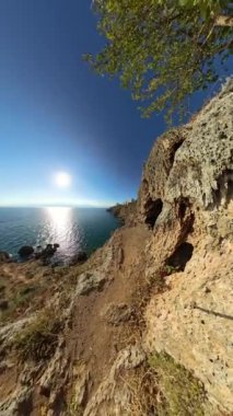 Dikey video. 360 derece dönüş. Geniş Açı, engebeli bir sahil uçurumu, denizin altında uzandığı bir yol. Güneş ışıl ışıl parlıyor, kayalık oluşumlara ışık tutuyor ve dramatik ve maceralı bir kıyı yolu oluşturuyor..