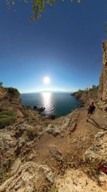 Dikey video. Onu seyreden yalnız bir yürüyüşçü, güneşli bir sahil yolunda yürüyor. Deniz parlak güneşin altında parıldıyor. Engebeli uçurumlar ve geniş okyanus manzarası huzurlu ve geniş bir manzara yaratır..