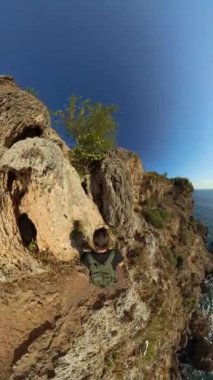 Dikey video. Geniş Açı. Bir dağcı okyanusun yukarısındaki engebeli bir uçuruma tırmanır. Parlak güneş pürüzlü kayaların üzerine ışık saçar. Sahne, kıyı keşiflerinin maceracı ruhunu yakalıyor..