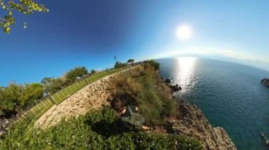 Bir adam uçurumun kenarındaki bir çitten denize doğru tırmanıyor. Sahildeki bir uçurumun tepesindeki çimenli bir parkın manzaralı geniş açılı görüntüsü, denize bakıyor. Parlak güneş ve geniş okyanus ufku barışçıl ve güzel bir doğal manzara yaratır..