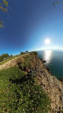 Dikey video. Bir adam uçurumun kenarındaki bir çitten denize doğru tırmanıyor. Sahildeki bir uçurumun tepesindeki çimenli bir parkın manzaralı geniş açılı görüntüsü, denize bakıyor. Parlak güneş ve geniş okyanus ufku barışçıl ve güzel bir doğal manzara yaratır..