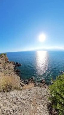 Dikey video. Uzaklaş, yakınlaş. Önünde engebeli kıyı kayalıkları olan güneşli okyanusun panoramik manzarası. Sakin mavi gökyüzü ve sakin deniz huzurlu ve geniş bir kıyı manzarası yaratır..