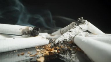 Yansıtıcı bir yüzeye yayılmış sigara ve küllerin yükselen dumanla geniş bir görüntüsü. Sahne, sigara kalıntılarını düşük açılı perspektifte gösteriyor..