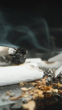 Dikey video. Yansıtıcı bir yüzeye yayılmış sigara ve küllerin yükselen dumanla geniş bir görüntüsü. Sahne, sigara kalıntılarını düşük açılı perspektifte gösteriyor..