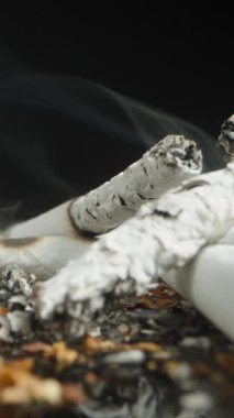 Dikey video. Yüzeyde yanmış ve buruşmuş sigara izmaritleri ve arka planda yükselen dumanla dolu bir yakınlaştırma çekimi. Dinamik perspektif, atılmış sigara ve küller üzerinde yoğun bir odak oluşturuyor..