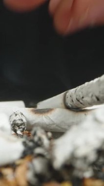 Vertical video. A close-up of a hand extinguishing a cigarette butt in a pile of ashes, with smoke rising from the remnants. The detailed view highlights the act of putting out the cigarette.