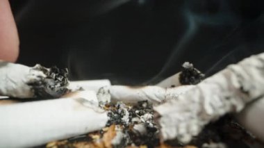 Close-up shot of a hand putting out a cigarette butt in a pile of ashes, with rising smoke. The focus is on the fingers pressing the cigarette into the ash.