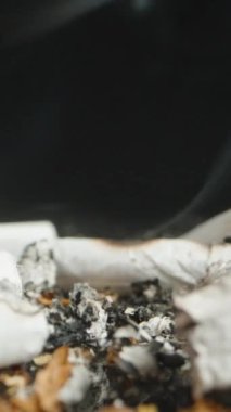 Vertical video. Close-up shot of a hand putting out a cigarette butt in a pile of ashes, with rising smoke. The focus is on the fingers pressing the cigarette into the ash.