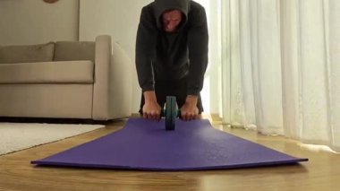 Bir adam, ahşap zeminli parlak bir odada mor bir paspas üzerinde, dinamik bir odak noktası için ön alçak bir açıdan vurularak, bir kas silindirinin sapını sıkıca kavrıyor..