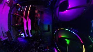 360 numaralı kamera çantanın içinde dönerken, canlı RGB aydınlatma ve soğutma vantilatörleri olan bir oyun bilgisayarının içinde yakın çekim, geniş açı görüntüsü. Tişörtsüz bir adam arka planda bir masada oturur loş bir odada cips yer..