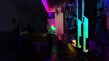 Geniş açı, bir oyun bilgisayarının içinden yakın çekim, parlayan RGB ışıkları ve donanım bileşenleri gösteriliyor. Loş arka planda, bir adam masada oturuyor, iki bacağı masanın üzerinde, karanlık bir odada rahat bir atmosfer yaratıyor..