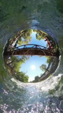 Dikey video. Nehir üzerindeki ahşap bir köprünün Tiny Planet görüntüsü, yemyeşil ağaçlar ve mavi gökyüzü ile çevrili, çevreyi bir daireye dönüştüren küresel bir panorama etkisi yaratıyor..
