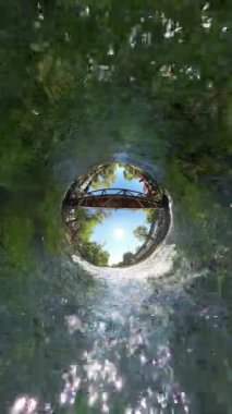 Dikey video. Kamera, Tiny Planet etkisinden geniş açılı bir görüntüye geçiyor. Nehrin ağaçlar ve yeşilliklerle çevrili ahşap bir köprünün altından akışını çekiyor. Güneş ışığı sudan yansıyor..