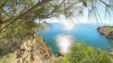 Kıyı kayalıklarının geniş açılı bir görüntüsü, yeşillik engin mavi okyanus manzarasını çerçeveliyor, ve güneş suların yüzeyine parlak bir şekilde yansıyor, barışçıl bir deniz manzarası yaratıyor..