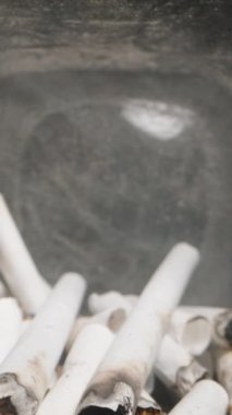 Dikey video. Dolly Slider, kapat. Sigara izmaritleriyle dolu bir cam kavanozun içinde, sigara içmenin sağlık risklerini ve çevresel etkilerini, görünür külleri ve yanmış filtreleri, kapalı, kirli bir alanda vurguluyor..