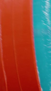 Dikey video. Eşsiz kırmızı ve mavi bir deodorant çubuğuna yakın plan, merkezi deliği ve pürüzsüz dokuyu vurguluyor, ürünün işlevsel ve estetik yönlerine odaklanıyor.