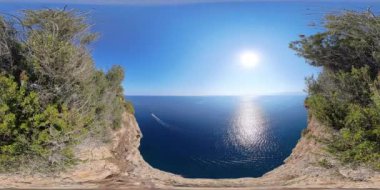Uçurumdan 360 derecelik panoramik bir manzara, engin okyanusa tepeden bakan, üzerinde güneş ışıl ışıl parlayan, sahnenin her iki tarafındaki yemyeşil yapraklarla çerçevelenmiş.