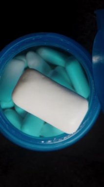 Dikey video. Yavaş çekimde, bir el, sakız parçalarıyla dolu açık mavi bir kapta düşmekte olan nane şekerli sakızı yakalar. Detaylı olarak hassasiyet ve tazeliği vurgular..