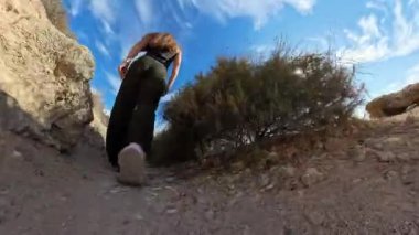 Uçurumun kenarındaki engebeli bir yolda yürüyen bir kadının, parlak mavi gökyüzünün altında, kuru bitki örtüsüyle çevrili, yakın açılı bir çekimi.