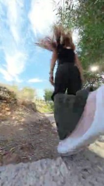 Dikey video. Doğal bitki örtüsüyle çevrili basamakları tırmanan bir kadının alçak açılı görüntüsü, açık mavi bir gökyüzü ve engebeli bir uçurum yolu ile çevrelenmiş.