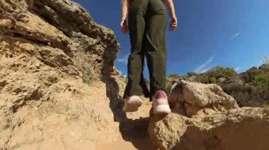 Açık mavi gökyüzünün altında kayalık bir yola tırmanan bir kadının alçak açılı çekimi, hareketi ve engebeli doğal manzarayı vurguluyor.